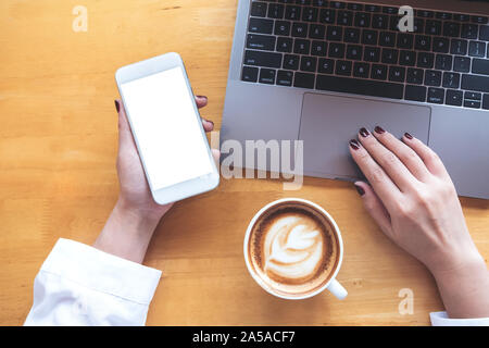Ansicht von oben mockup Bild der Hand einer Frau mit Mobiltelefon mit leeren weißen Bildschirm, während Sie laptop mit Kaffeetasse auf hölzernen Tisch Stockfoto