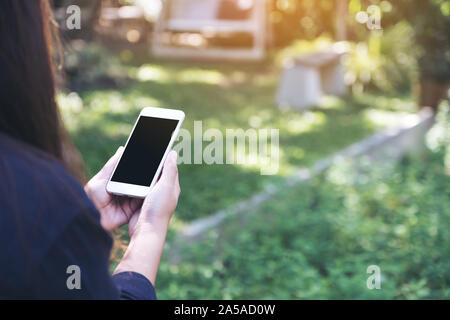 Mockup Bild von einer Frau mit und die Verwendung von weißen smart phone mit leeren schwarzen Bildschirm im Außenbereich mit grünen Natur Hintergrund Stockfoto