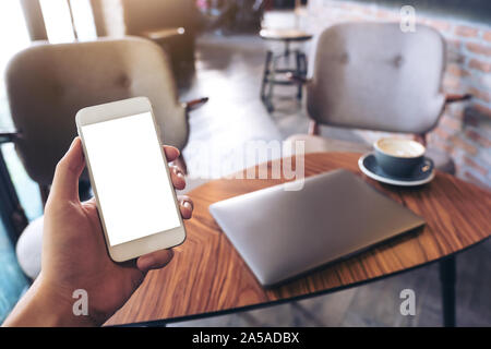 Mockup Bild der Hände ist ein Mann mit weißer Handy mit leeren Desktop, Laptop und Kaffeetasse in der modernen Loft Cafe Stockfoto