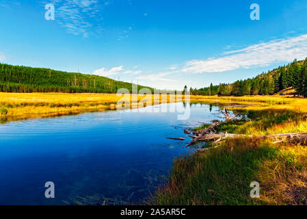 Ruhigen blauen Fluss durch Golden grasbewachsenen Senke mit grün bewaldeten Bergen unter einem blauen Himmel mit weißen Wolken. Stockfoto