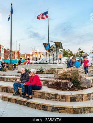 HICKORY, NC, USA-11 OCT 2019: Seniorenpaar sitzt auf einer Treppe auf dem Stadtplatz, mit Kindern spielen auf ausgestellten Kanonen und Fahnen im Hintergrund. Stockfoto