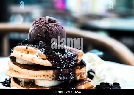 Eine Nahaufnahme von dekadenten Buttermilchpfannkuchen mit geschnittenen Bananen, reichhaltigem Schokoladensirup, Schokoladenchips und Eis. Stockfoto