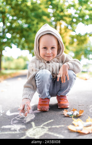 Kleiner Junge, 4-5 Jahre alt, zeichnet mit Buntstiften auf Bürgersteig, in warmen Pullover mit Kapuze, glücklich, Lächeln, genießt Wochenende. Im Sommer, im Herbst Park Stockfoto