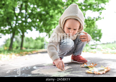 Kleiner Junge, 4-5 Jahre alt, zeichnet mit Buntstiften auf Bürgersteig, in warmen Pullover mit Kapuze, schafft kreative Zeichnungen, im Sommer, im Herbst Park. Hintergrund Stockfoto