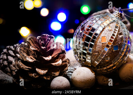 Weihnachtsschmuck Szene mit Lichterketten vor einem schwarzen Hintergrund. Eine silberne Christbaumkugel mit Spiegel sitzt neben zwei Tannen Zapfen auf einem Bett aus Kieselsteinen. Stockfoto