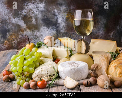 Essen still life: Verschiedene köstliche Käsesorten, Glas Weißwein, Nüssen, Obst, würzige Kräuter und Zwiebeln baguette auf einem urigen Hintergrund. Stockfoto