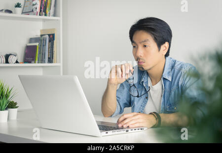 Asiatische Casual Unternehmer oder Freiberufler in Denim oder Jeans Shirt halten Brillen und Arbeiten mit Laptop im Home Office. Ernsthafte casual Geschäftsmann oder F Stockfoto