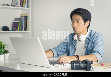 Asiatische freiberufliche Videofilmer in Denim oder Jeans Hemd bearbeiten Multimedia Datei per Laptop im Home Office. Freiberufliche Videofilmer arbeiten mit Technologie Stockfoto