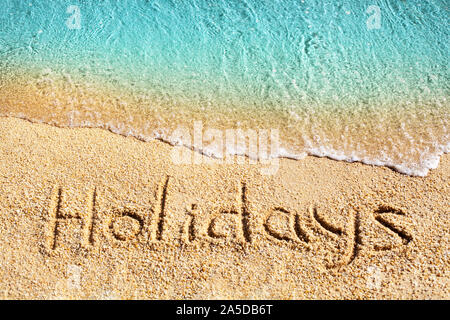 Wort Urlaub auf gelbem Sand geschrieben, blaues Meer, weißer Schaum Top View close up, Handschrift schreiben Text, Meer Wasser, Sommer Ferien Konzept Stockfoto