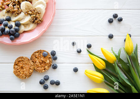 Weißer Tisch serviert, ein Teller Banane Stücke, Heidelbeere und Haferflocken Cookies. Gelbe tupic Blumen Dekorationen. Stockfoto