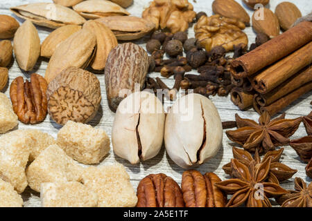 Sortiment von Nüssen und Gewürzen, Mix aus verschiedenen Arten von Nüssen, wie Nussbaum, Muskatnuss, Pecan und Mandel. Zimtstangen und braunen Zucker auf Holz grau Bac Stockfoto