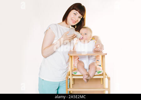 Eine junge schöne Mutter Feeds ein Baby aus einer Flasche. Das Kind sitzt auf einem Stuhl, der auf einem weißen Hintergrund. Stockfoto