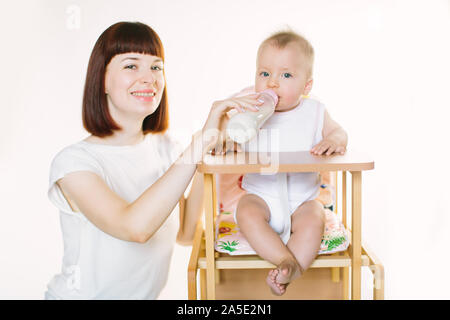 Eine junge schöne Mutter Feeds ein Baby aus einer Flasche. Das Kind sitzt auf einem Stuhl, der auf einem weißen Hintergrund. Stockfoto