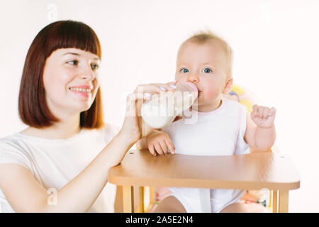 Eine junge schöne Mutter Feeds ein Baby aus einer Flasche. Das Kind sitzt auf einem Stuhl, der auf einem weißen Hintergrund. Stockfoto