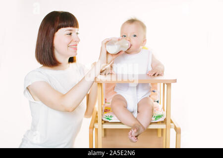 Eine junge schöne Mutter Feeds ein Baby aus einer Flasche. Das Kind sitzt auf einem Stuhl, der auf einem weißen Hintergrund. Stockfoto