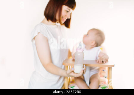 Eine junge schöne Mutter Feeds ein Baby aus einer Flasche. Das Kind sitzt auf einem Stuhl, der auf einem weißen Hintergrund. Stockfoto