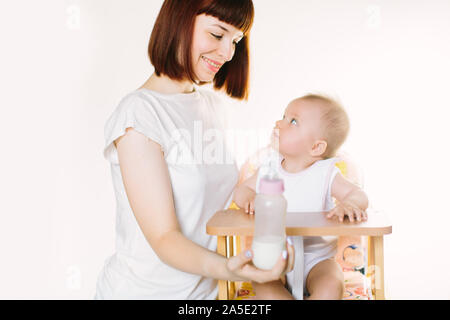 Eine junge schöne Mutter Feeds ein Baby aus einer Flasche. Das Kind sitzt auf einem Stuhl, der auf einem weißen Hintergrund. Stockfoto