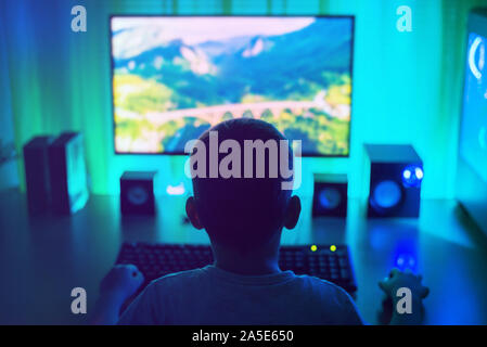 Jungen Arbeiten am Computer. Dunkle Szene mit viel Licht. Ansicht von hinten, dritter Person. Konzept eines jungen Hackers. Stockfoto