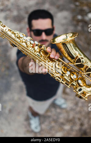 Junger Mann mit Bart und Sonnenbrille Holding eine glänzende Saxophon vor seinem Gesicht im Freien statt. Saxophonist mit einem Saxophon, blur Effekt. Stockfoto