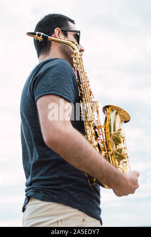 Junger Mann mit Bär und Sonnenbrille mit einem Saxophon mit bewölktem Himmel Hintergrund. Saxophonist und weg schauen an einem bewölkten Tag. Stockfoto