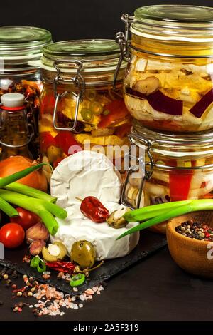 Eingelegter Käse mit roten Zwiebeln. Hausgemachte Zubereitung von eingelegtem Käse. Gebeizt essen. Glas mit Camembert, mariniert in Öl auf schwarzen Holztisch. Stockfoto