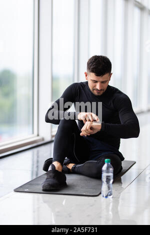 Ruhezeit. Zuversichtlich muskulösen jungen Mann tragen Sportkleidung und sitzen auf dem Boden mit Flasche Wasser im Loft Interieur Stockfoto