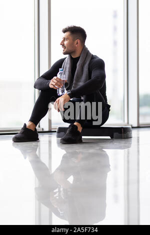 Ruhezeit. Zuversichtlich muskulösen jungen Mann tragen Sportkleidung und sitzen auf dem Boden mit Flasche Wasser im Loft Interieur Stockfoto