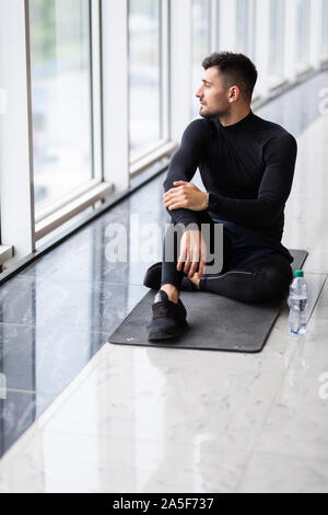 Ruhezeit. Zuversichtlich muskulösen jungen Mann tragen Sportkleidung und sitzen auf dem Boden mit Flasche Wasser im Loft Interieur Stockfoto