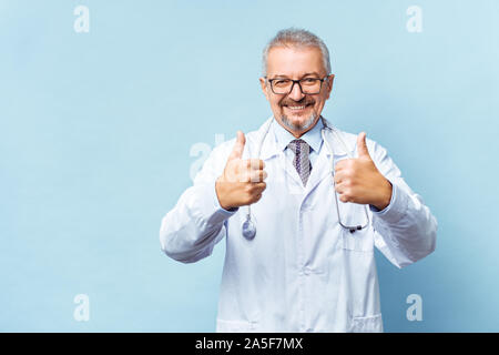 Lächelnd medizinische leitender Arzt mit einem Stethoskop. Auf einem blauen Hintergrund. Medic zeigt Daumen angehoben. Das Konzept der Menschheit den Sieg über die Krankheit Stockfoto