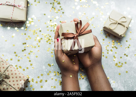 Schwarze Mädchen gibt ein Geschenk mit ihren eigenen Händen gemacht. Hände halten ein Handwerk Geschenk mit einem braunen Band auf einem grauen Hintergrund mit ein paar kleine Geschenke. Flach. Stockfoto