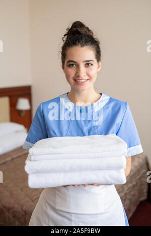 Junge fröhliche brunette Zimmermädchen mit Stack von sauberen, weißen weiche Handtücher im Zimmer stehend Stockfoto