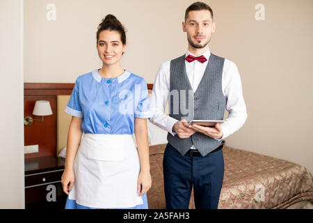 Glückliche junge Zimmermädchen in Uniform und elegante Porter mit Touchpad stand vor der Kamera Stockfoto