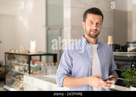 Hübscher Kerl in der casualwear Scrollen in Smartphones, während Sie auf der Suche nach Hintergrund der Cafe innen Stockfoto