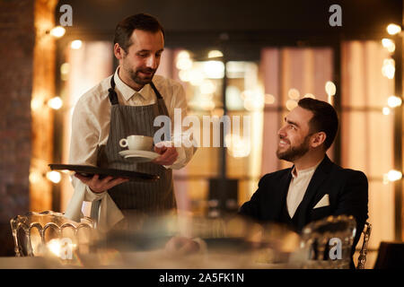 Seitenansicht Portrait von gut aussehenden bärtigen Geschäftsmann im Gespräch mit Kellner Kaffee bringen und glücklich lächelnd in Luxus Restaurant Stockfoto