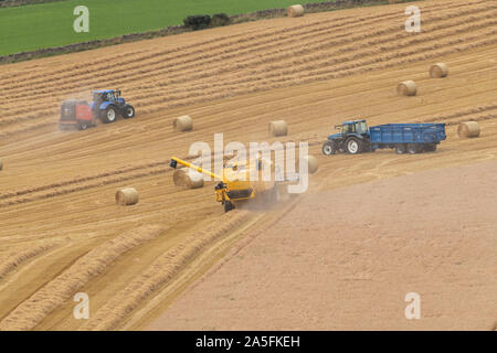 Ein Mähdrescher ernten Traktor bergauf, während ein Ballen Stroh und ein zweiter Traktor, wartet mit einem Anhänger in einem Feld in Aberdeenshire, Schottland Stockfoto