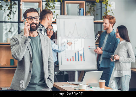 Startupers zusammen arbeiten im Büro bärtigen Mann in Gläsern close-up sprechen auf dem Smartphone heiter während Kollegen diskutieren Daten an Bord Stockfoto