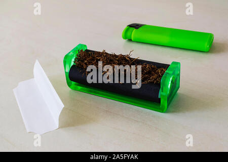 Grüne zigarette Walzmaschine mit Tabak, grüne leichter und Zigarettenpapier auf einem weißen Tisch. Die Zigaretten mit Pfeifentabak zu Hause. Stockfoto