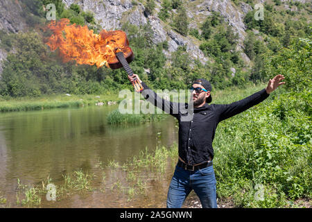 23 Jahre alter Mann mit Bart in einem schwarzen T-Shirt, eine Kappe und Sonnenbrille hält eine brennende Gitarre. Vor dem Hintergrund blauer Himmel und die Berge. heavy metal gui Stockfoto