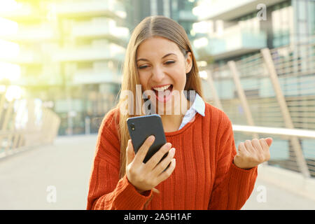 Aufgeregt gewinnende Frau gerade ihre smart phone Outdoor. Euphorisch fröhliche Frau ihr smart phone Kontrolle in der modernen Stadt. Stockfoto