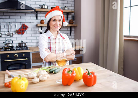 Positiv glückliche Erwachsene Frau vor der Kamera posiert in der Küche. Festliche rot Hut tragen. Hände gekreuzt. Silvester oder Weihnachten feiern. Allein Kochen Stockfoto