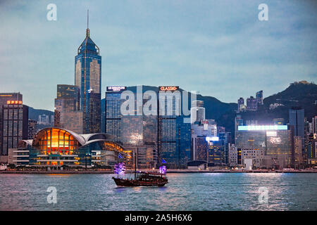 Traditionelles Trödelboot im Victoria Harbour, das an den Gebäuden der Central Waterfront vorbeifährt, die bei Sonnenuntergang beleuchtet sind. Hongkong, China. Stockfoto