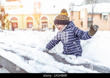 Kleiner Junge, 3-5 Jahre alt, spielt mit Schnee im Winter auf der Straße, in warme Mütze blauen Overalls, Hintergrund Der schneewehen von Schnee. Emotionen Spaß entspannten Spaß Stockfoto