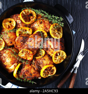 Hähnchen Schenkel mit gerösteten Zitronenscheiben und Thymian in eine schwarze Keramik holländischen Ofen auf einem Holztisch, horizontale Ansicht von oben, flatlay, Clo Stockfoto