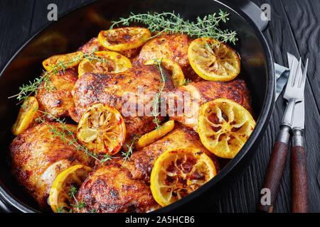In der Nähe von Hot Fried Chicken Schenkel mit gerösteten Zitronenscheiben und Thymian in eine schwarze Keramik Teller auf einem Holztisch, horizontale Ansicht von oben, flatla Stockfoto