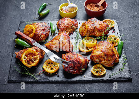 In der Nähe von knusprig gebratenen Hühnerkeule mit gerösteten Zitronenscheiben, jalapeno Pfeffer und Thymian auf einen schwarzen Stein Fach auf einer konkreten Tabelle, Ansicht von Abo Stockfoto