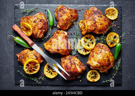 Knusprig gebratene Hühnerkeule mit gerösteten Zitronenscheiben und Thymian auf einen schwarzen Stein Board auf einer konkreten Tabelle, Ansicht von oben, flatlay, close-up Stockfoto