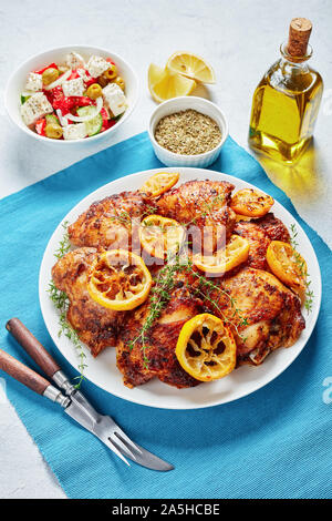 Knusprig gebratene saftige Hühnerkeule mit gerösteten Zitronenscheiben auf einer weißen Platte auf einer konkreten Tabelle mit griechischen Salat in eine Schüssel, Ansicht von oben Stockfoto
