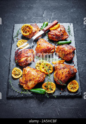 In der Nähe von knusprig gebratenen Hühnerkeule mit gerösteten Zitronenscheiben, jalapeno Pfeffer und Thymian auf einen schwarzen Stein Fach auf einer konkreten Tabelle, Ansicht von Abo Stockfoto