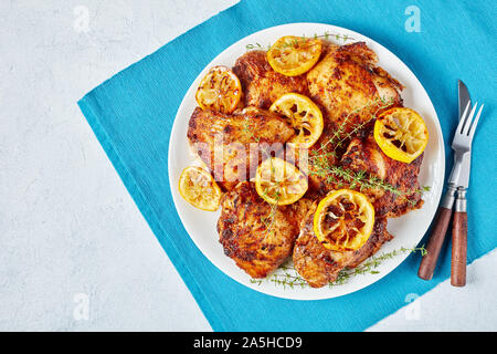 Knusprig gebratene saftige Hühnerkeule mit gerösteten Zitronenscheiben auf einer weißen Platte auf einer konkreten Tabelle, Flach, close-up Stockfoto