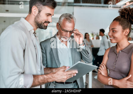 Lassen Sie mich sehen. Zuversichtlich fälligen Geschäfts Mann in formale Abnutzung auf digital Tablet im Stehen mit jungen Mitarbeiter im modernen Büro Stockfoto